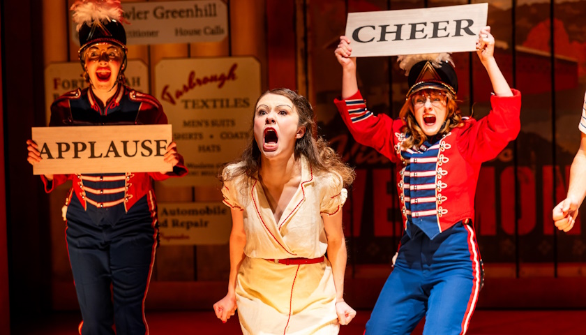 Three actors on stage, two hold signs saying "applause" and "cheer" while one screams