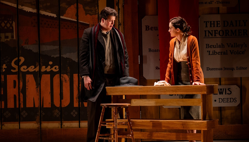 Two actors on stage at a table stand facing each other