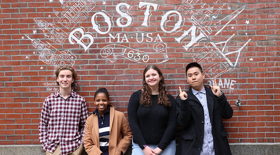 Four Suffolk students pose together in front of a brick wall that has a Boston-themed mural