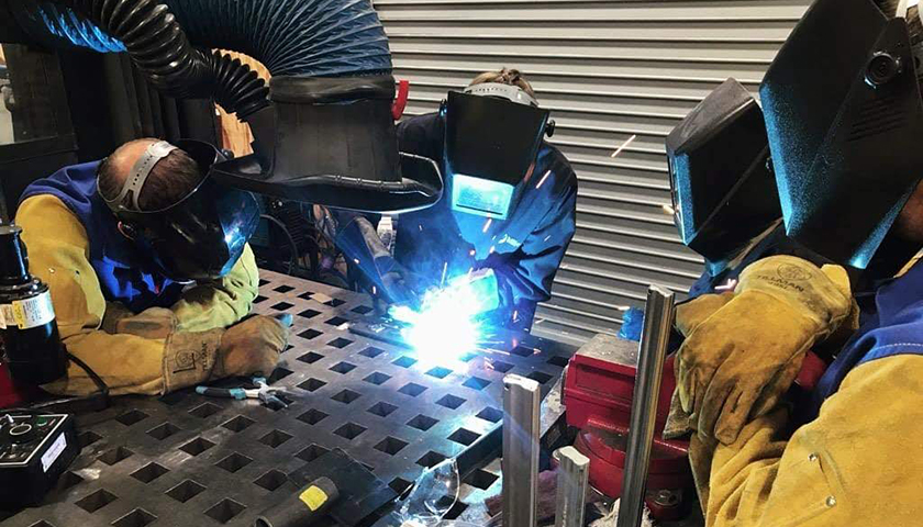 Three people in protective helmets and face coverings welding