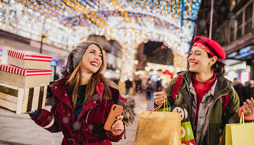 Image of two women holiday shopping, carrying bags and boxes, underneath an umbrella of lights