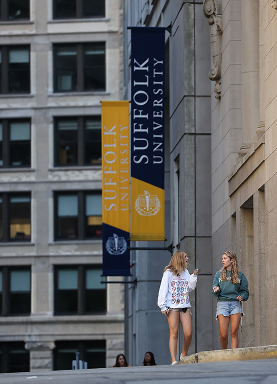 Suffolk students walk through campus, with tall buildings behind them.