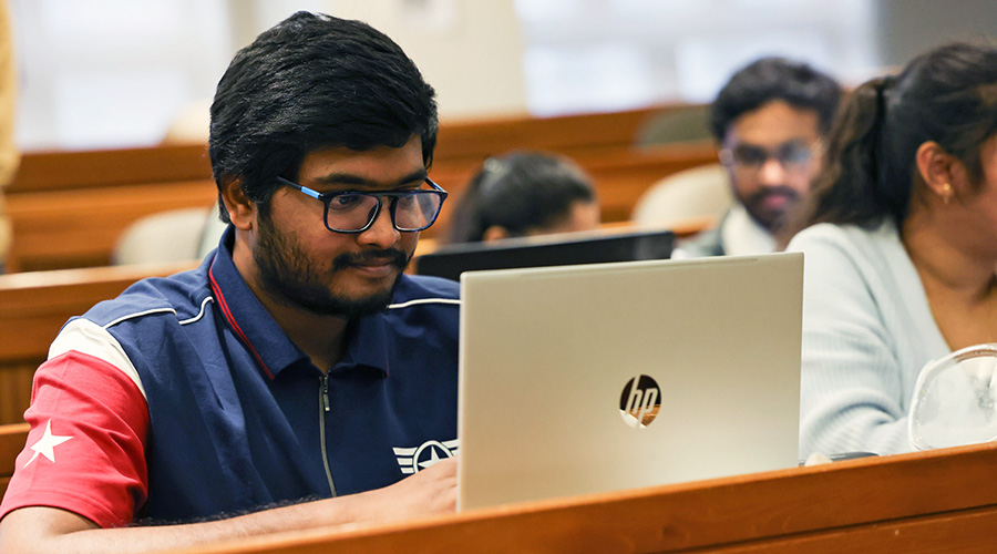 A student sitting at a bench desk focusing on his laptop in front of him.