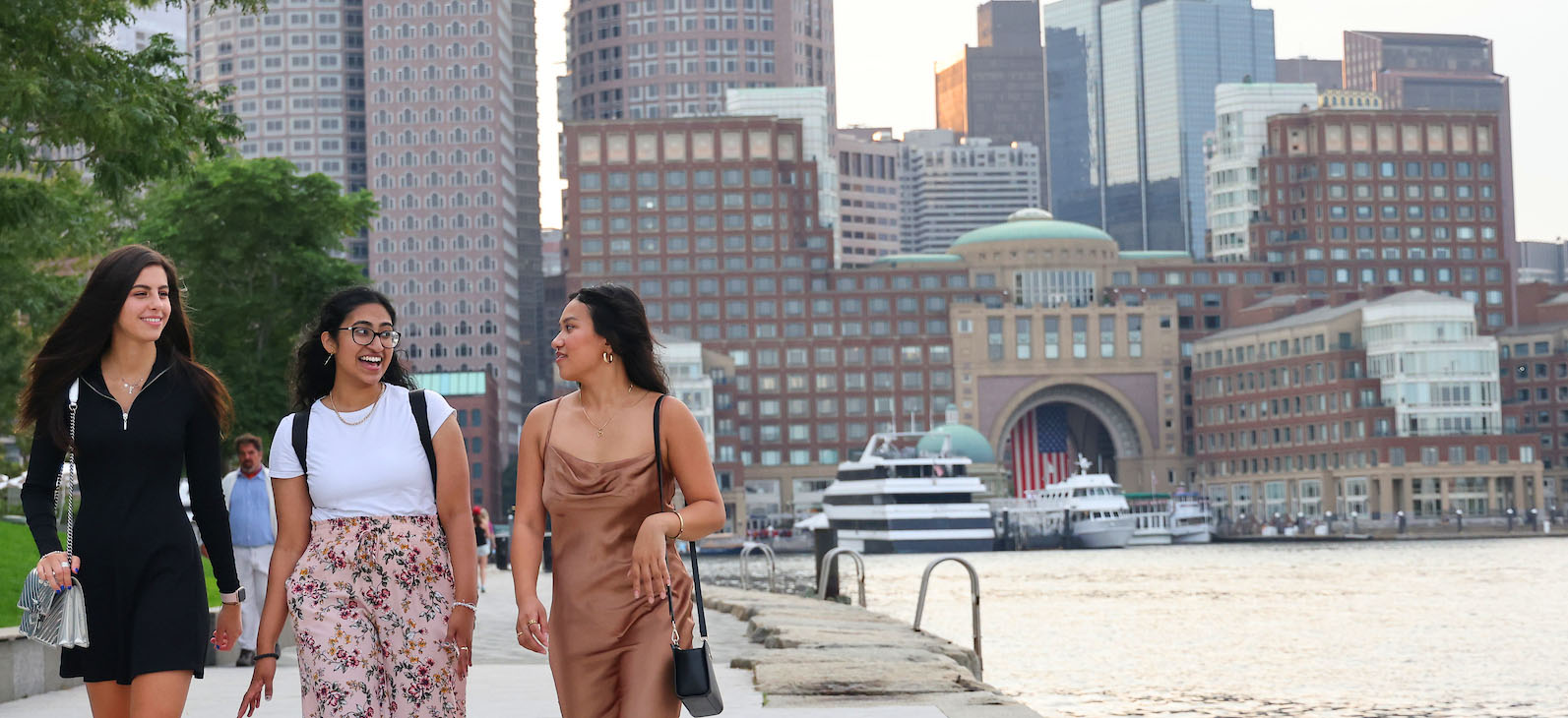 Three Suffolk students walk along the river with the city skyline behind them.