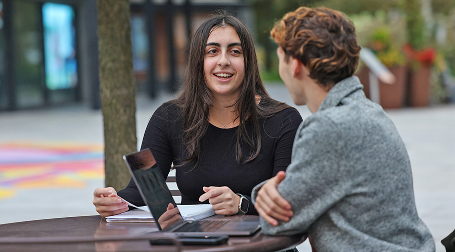 Two Suffolk students sitting at an outdoor table, one with a laptop and the other with a notebook, in conversation together.