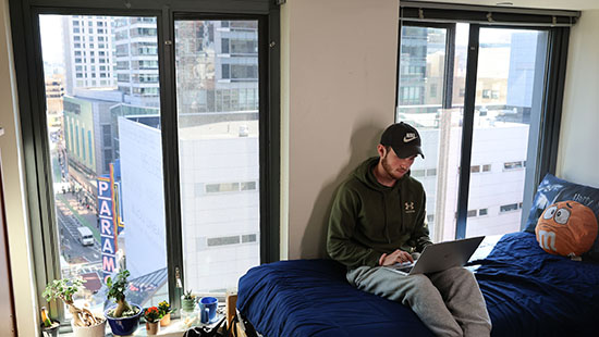 A student sits on the bed in his residence hall working on a laptop with Downtown Boston outside the window