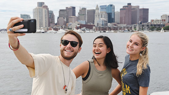 Three students take a selfie with the Boston skyline in the background