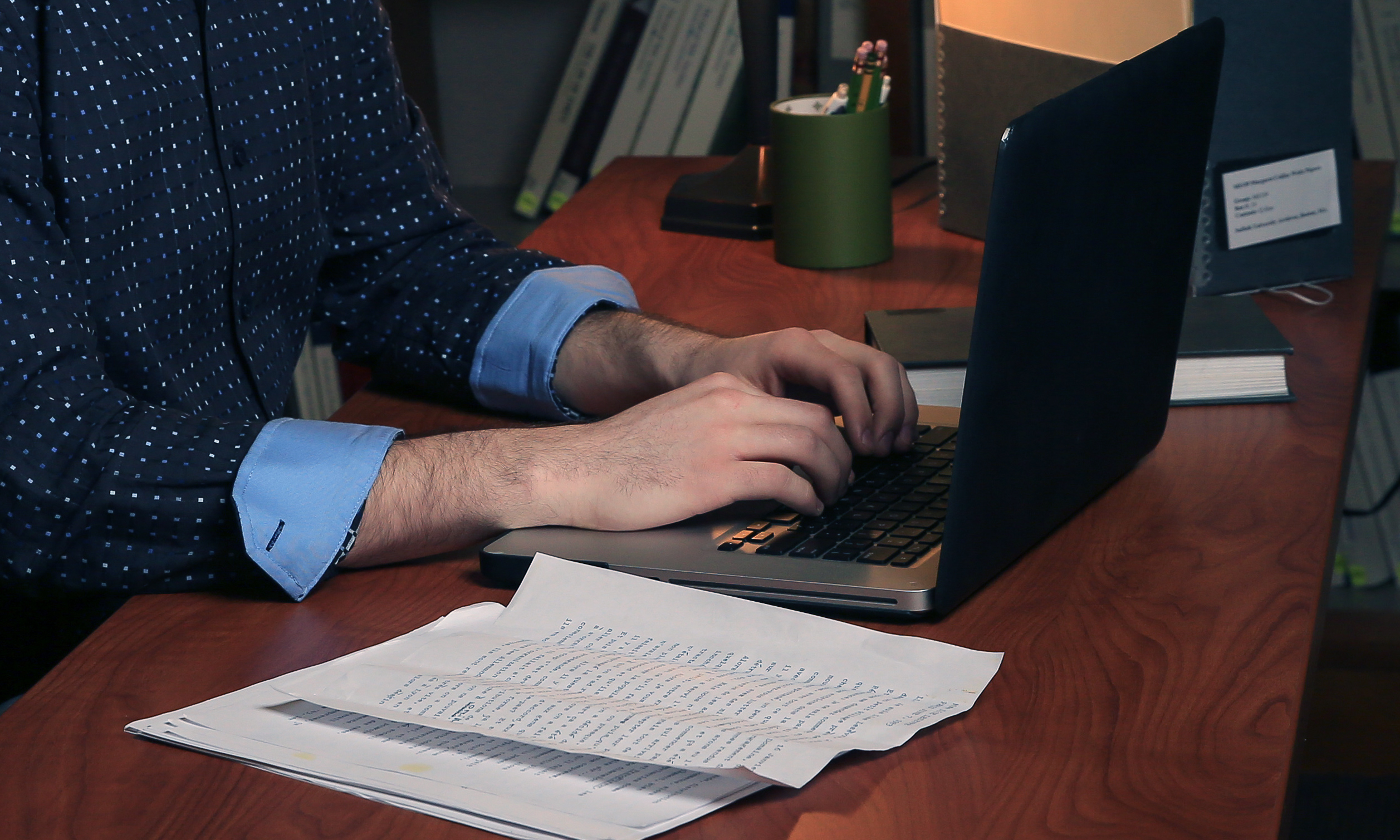 Someone working on a laptop with papers on the table next to them.