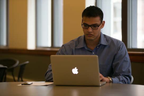 Student in blue collared shirt with square black glasses sits at a table and works on his laptop 