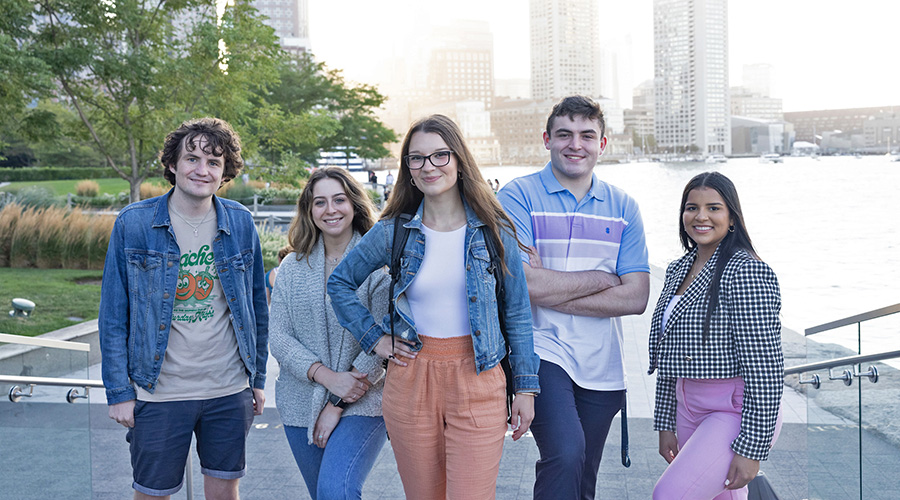 Five Suffolk students pose together along the riverfront with the Downtown cityscape behind them in the background.