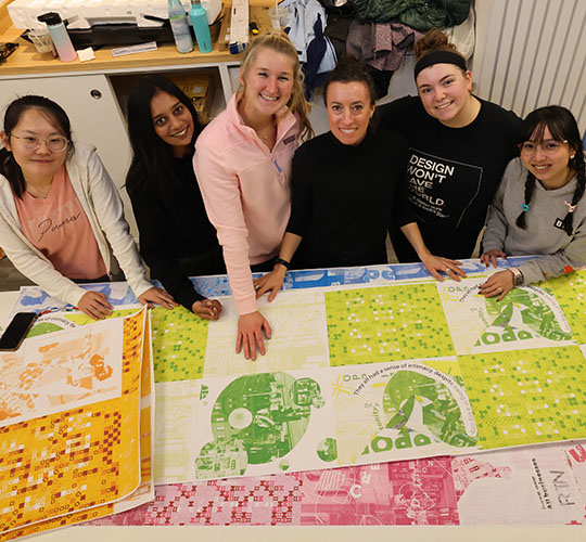 Several students geture at their artwork laid out on a table