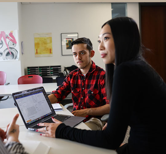 Two students at a desk working on a shared laptop