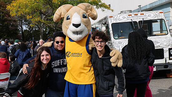 Three students take a picture with Suffolk ram mascot Rammy in front of a row of food trucks at an outdoor fall event.
