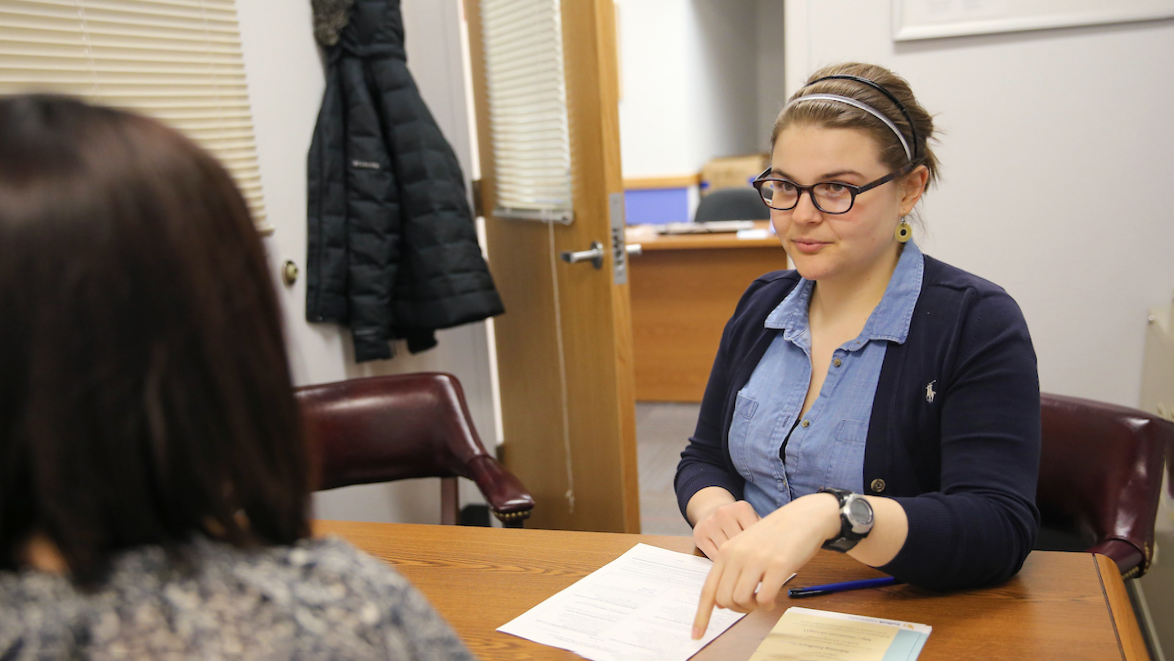 A student meets with an advisor, both sitting at a desk and the student points to a piece of paper on the desktop.