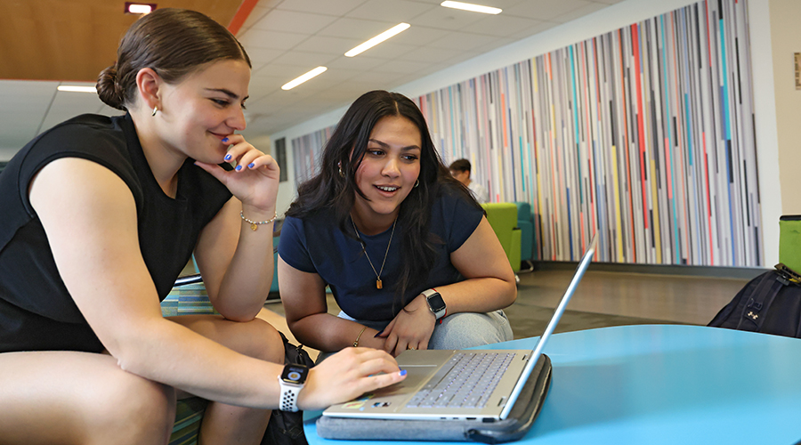 Two Suffolk students with engaged smiles sitting together in a lounge space on campus leaning over a shared laptop screen.