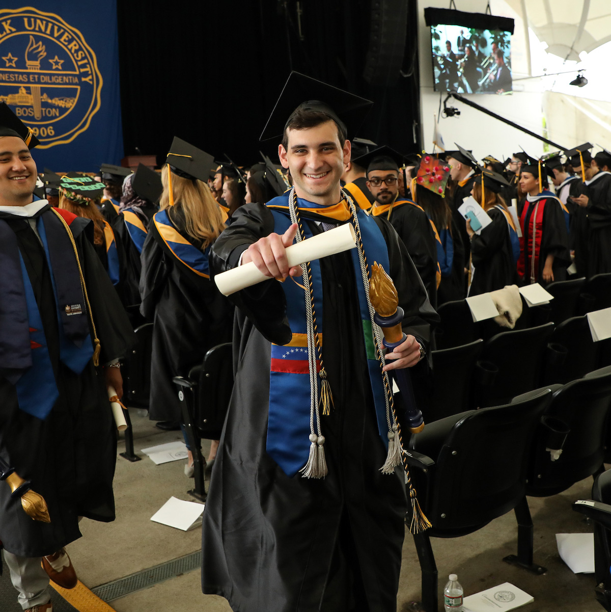 Dan smiling and point toward the camera while walking down the aisle in his cap and gown at Commencement.
