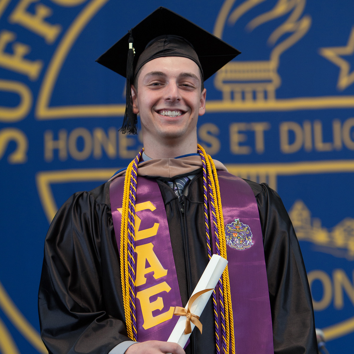 Dan holding up his scroll and smiling for the camera wearing his cap and gown while crossing the stage at Commencement.