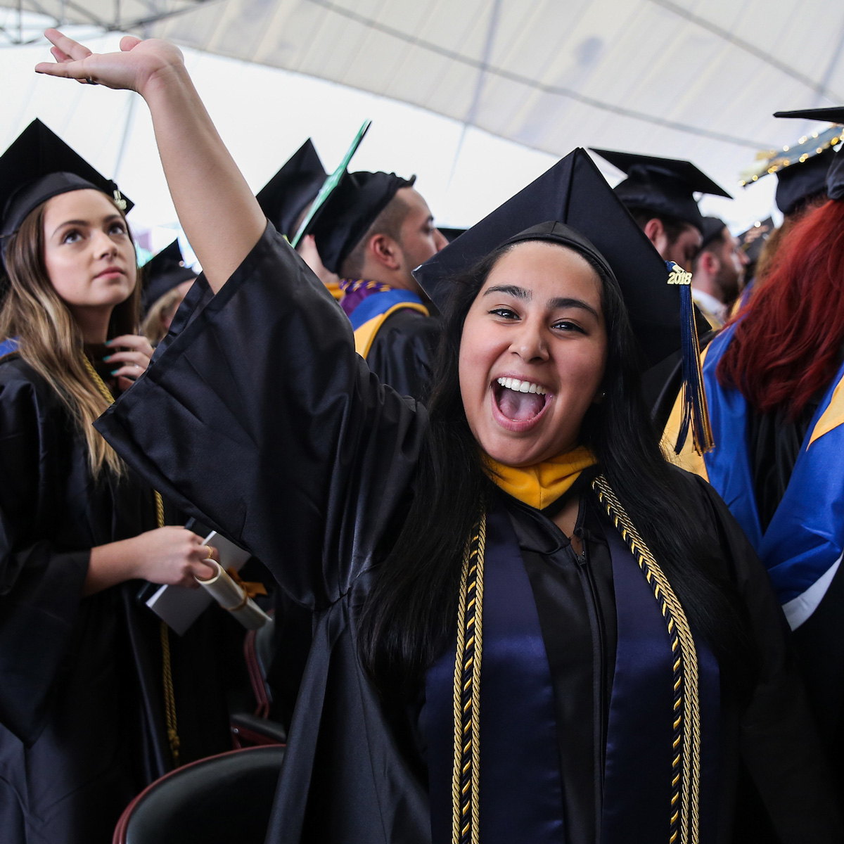 Grisell waving and smiling in the crowd wearing her cap and gown during Commencement.