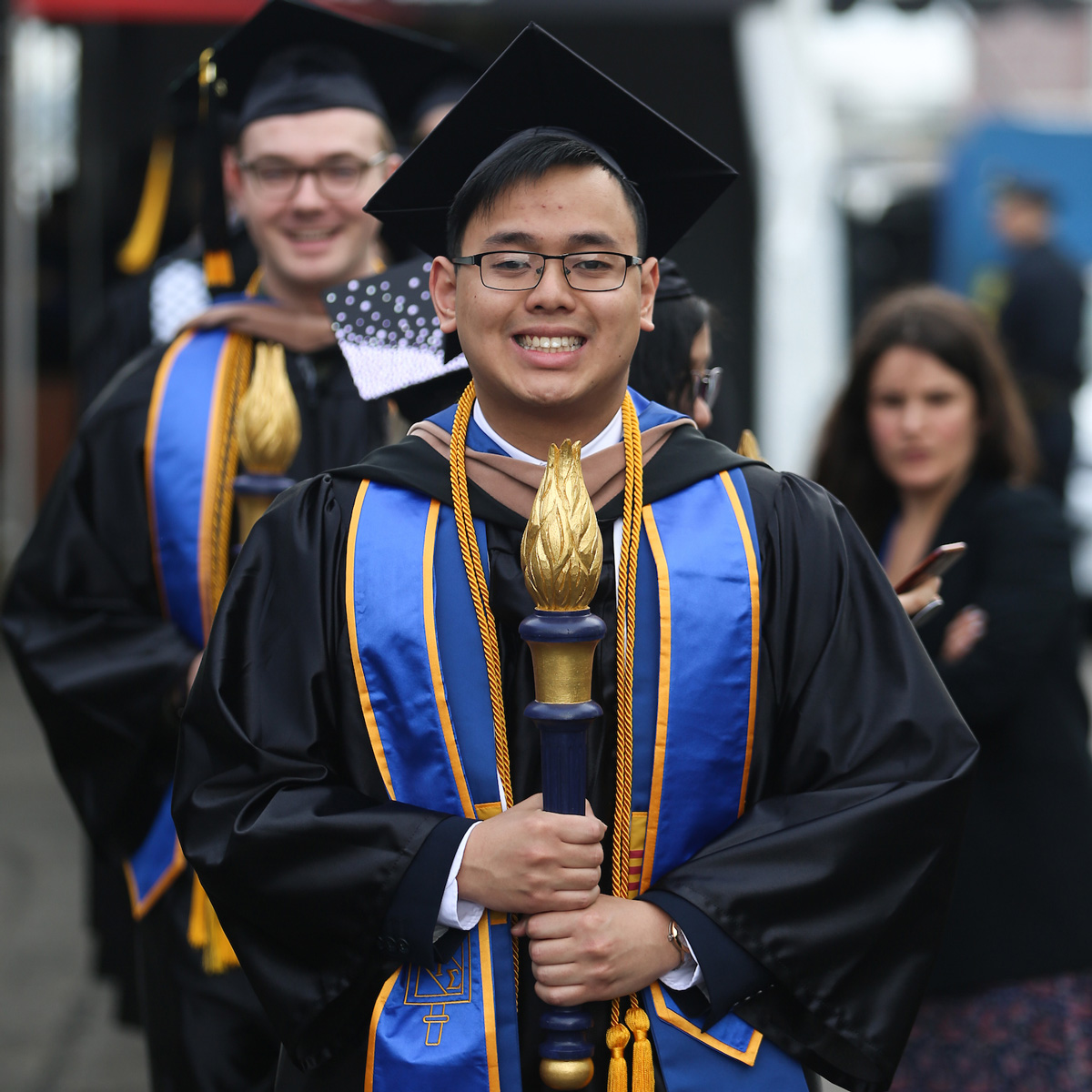 John carrying the torch leading the Commencement line wearing his cap and gown.