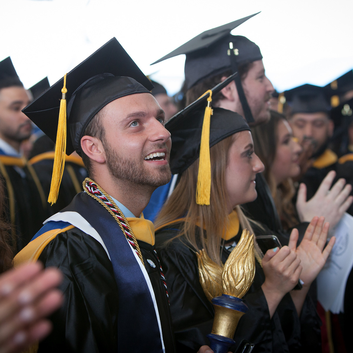 Jonathan holding the Suffolk torch in the crowd wearing his cap and gown during Commencement.