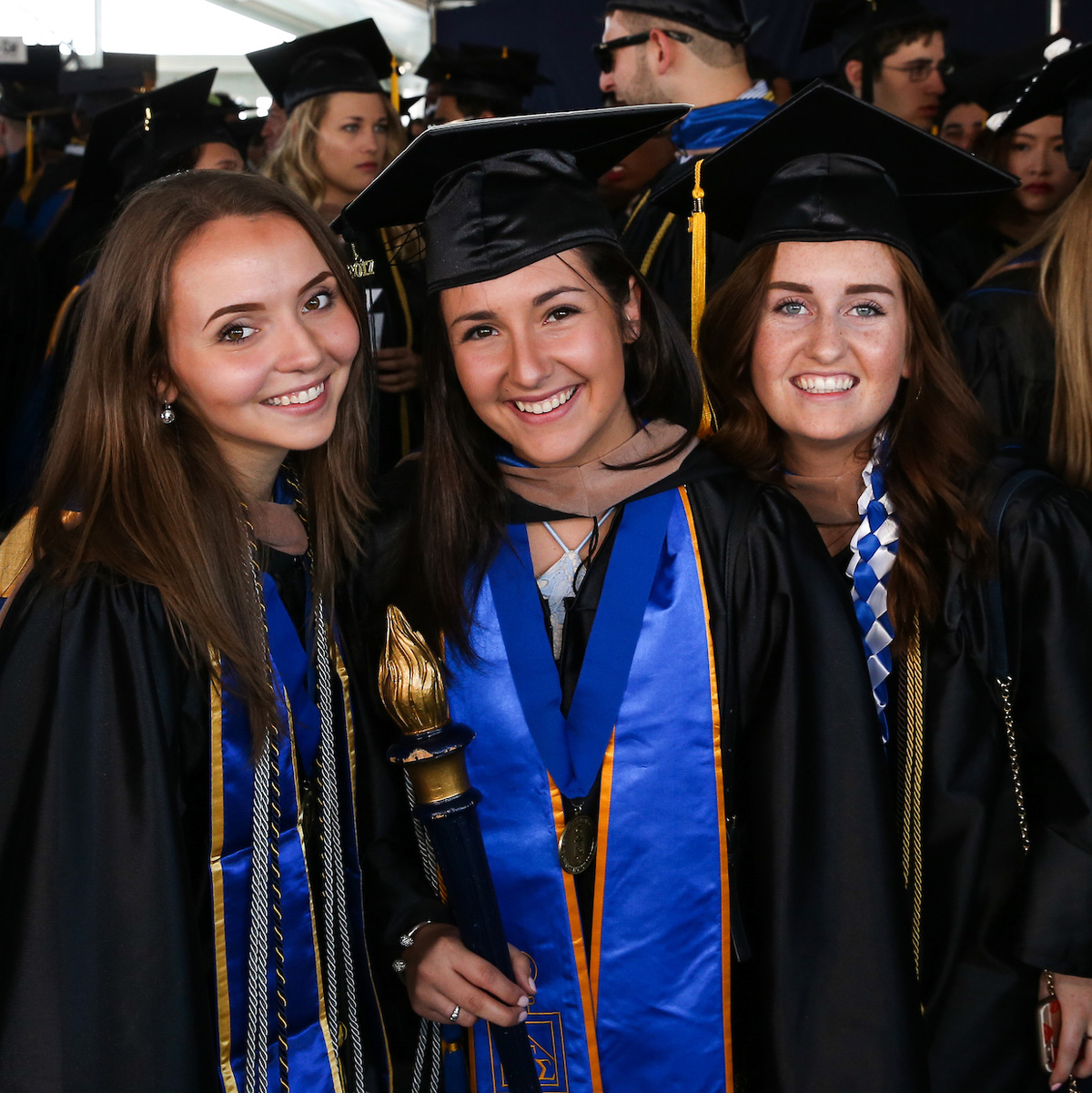 Sydney Fonseca carrying the Suffolk torch and posing in her cap and gown with classmates before Commencement.