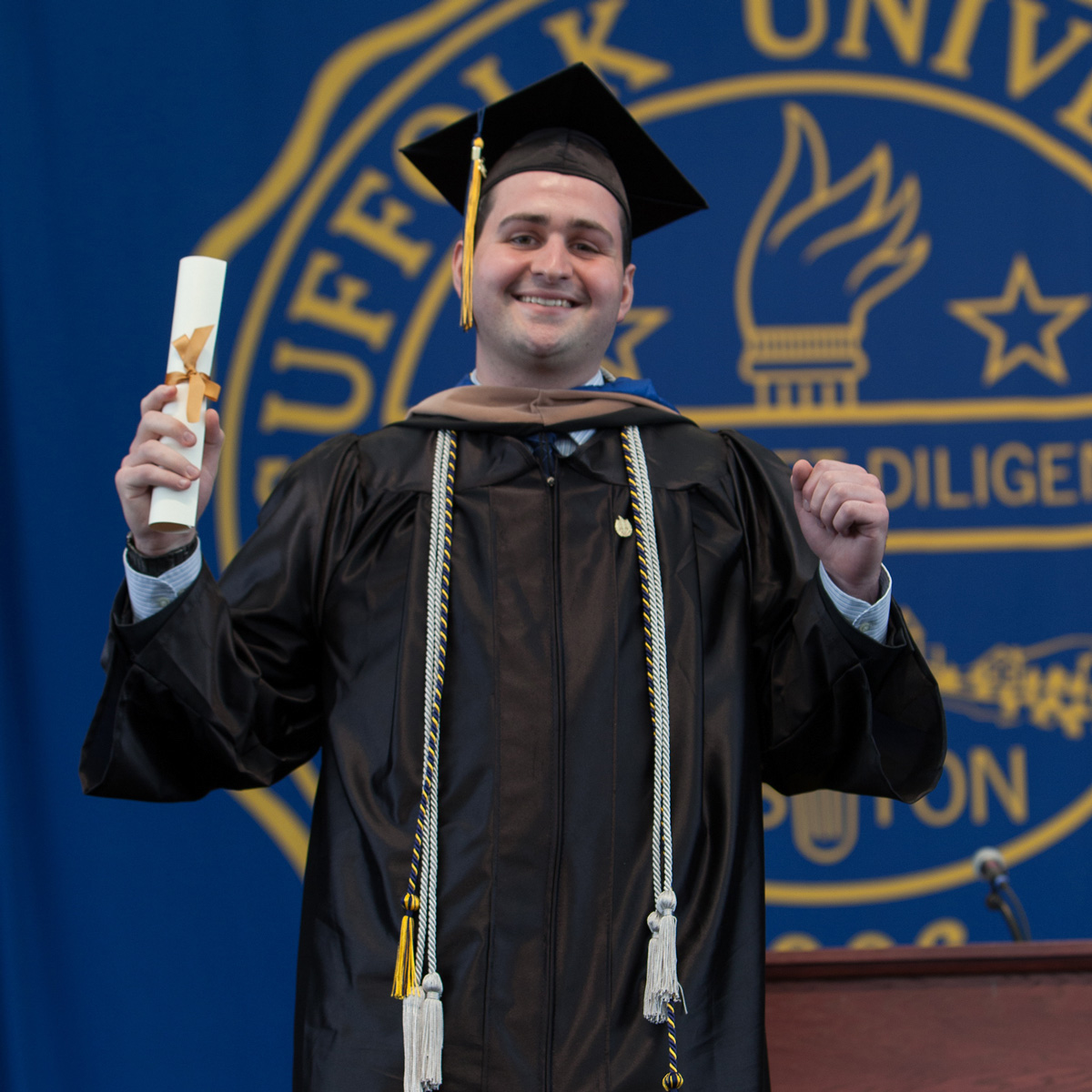Vincent LePore holding up his scroll wearing his cap and gown crossing the stage at Commencement.
