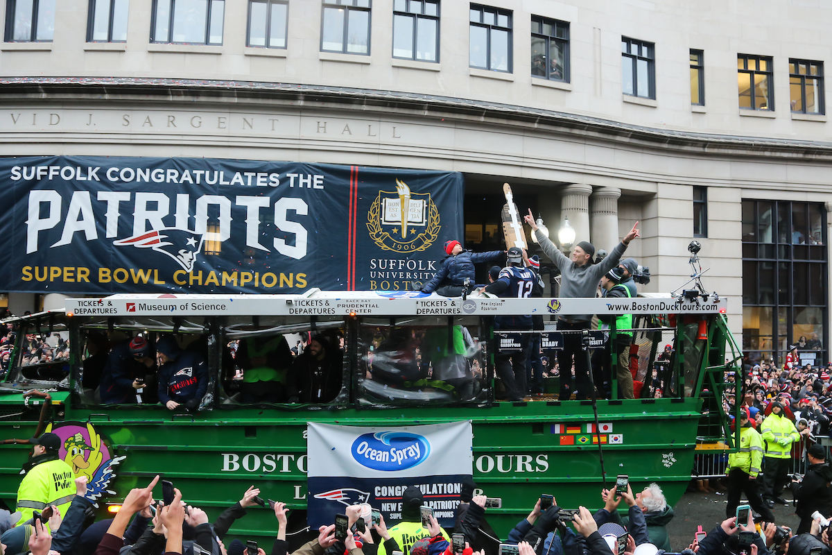 Quarterback Tom Brady raises his arms aboard a Duck boat rolling by Suffolk's congratulatory banner outside Sargent Hall.