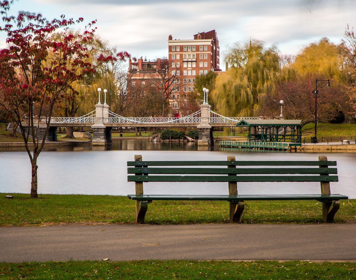 A park bench overlooking the Public Garden Suspension Bridge on a crisp, sunny autumn day.