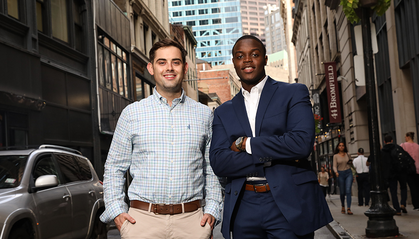 Mike Nay and Markendy Fils-Aime stand shoulder to shoulder outside with Downtown Boston buildings behind them