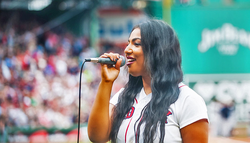 Rebecca Zama singing into microphone with crowd and Fenway's Green Monster in background