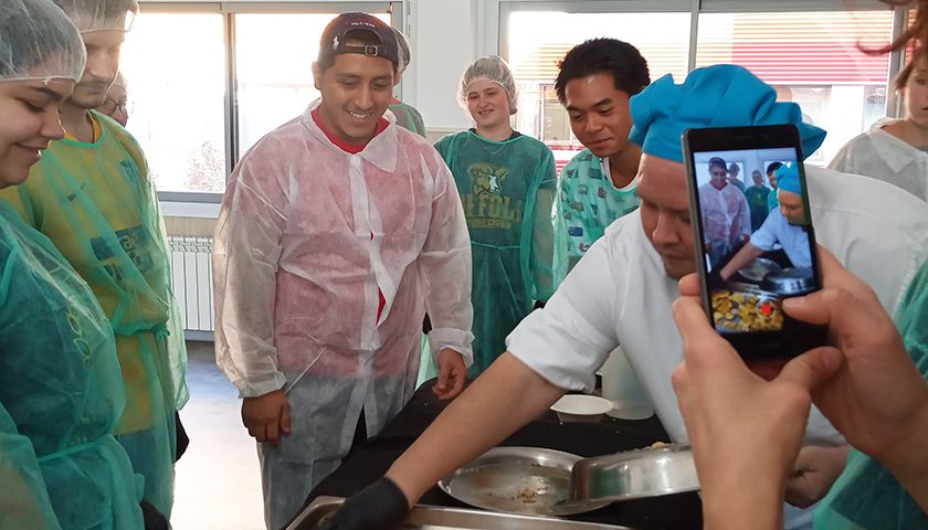 Students in shower caps and smocks help chef prepare food at refuge center