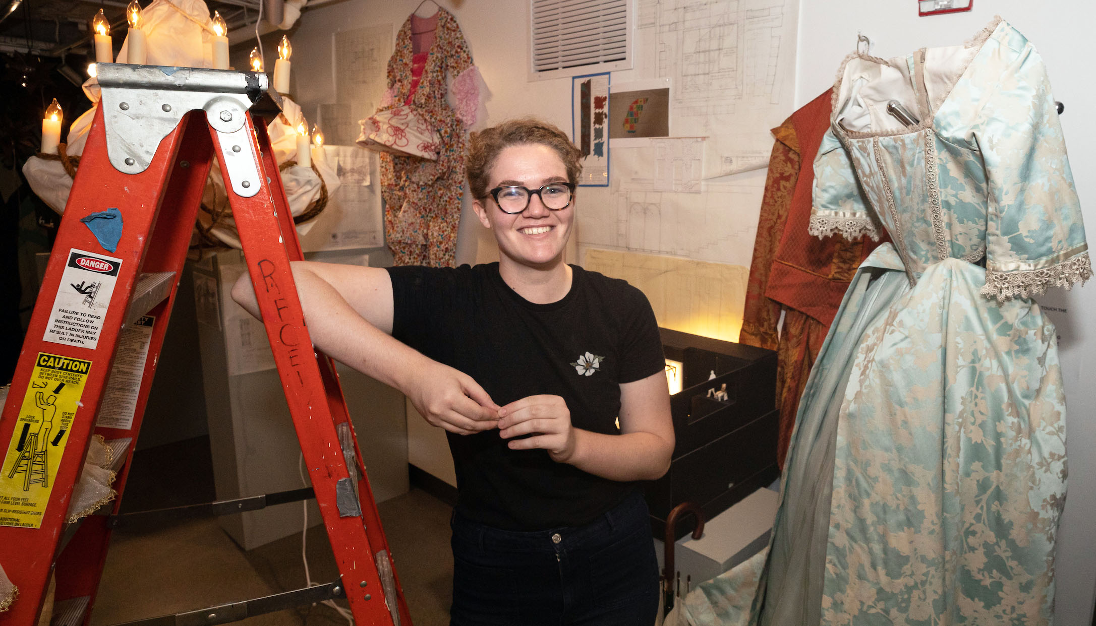 Micaleen Rogers stands with elbow resting on stepladder. Costume hangs on wall on stage right