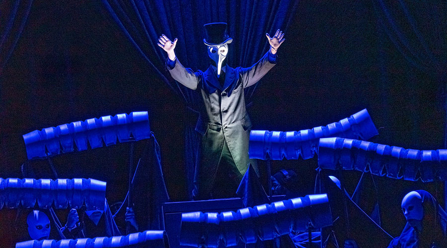 Man in white, beaked mask with arms upraised and surrounded by blue light