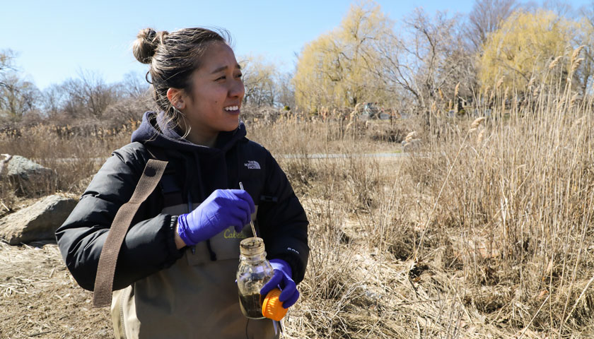 student stirs a soil and water mixture in a glass jar at the marsh