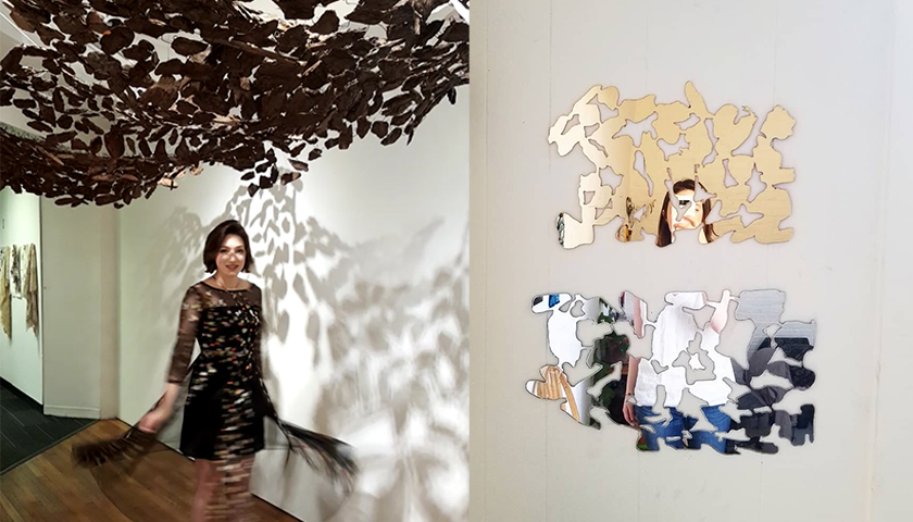 Woman standing below supended small pieces of bark, which throw shadows on wall; woman reflected in metallic shapes on wall