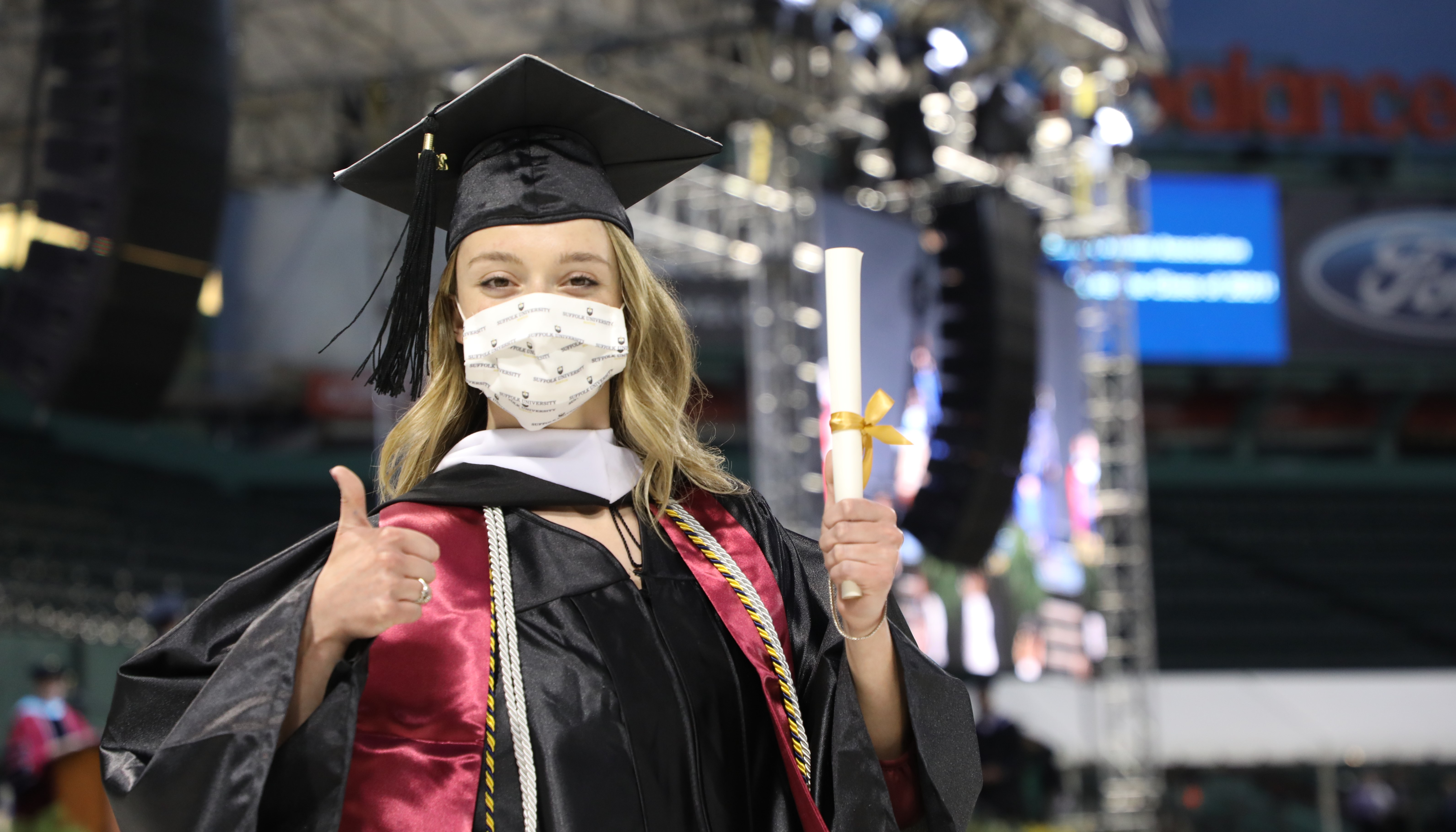 Student in a cap, gown, and mark gives a "thumbs up" sign as she holds her degree