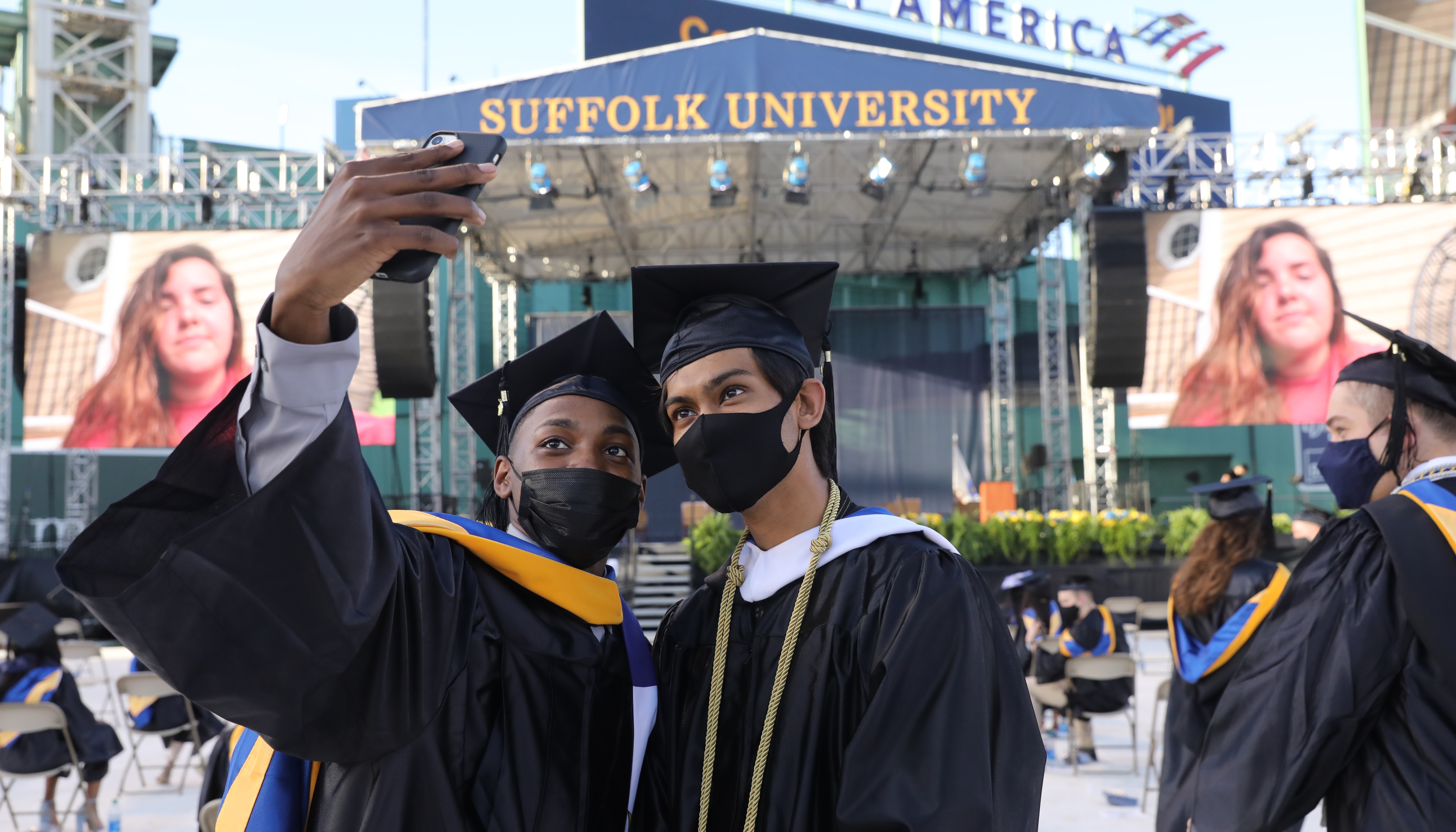 Two students pose in caps and gowns in front of the Class of 2020 Commencement stage as one takes a picture