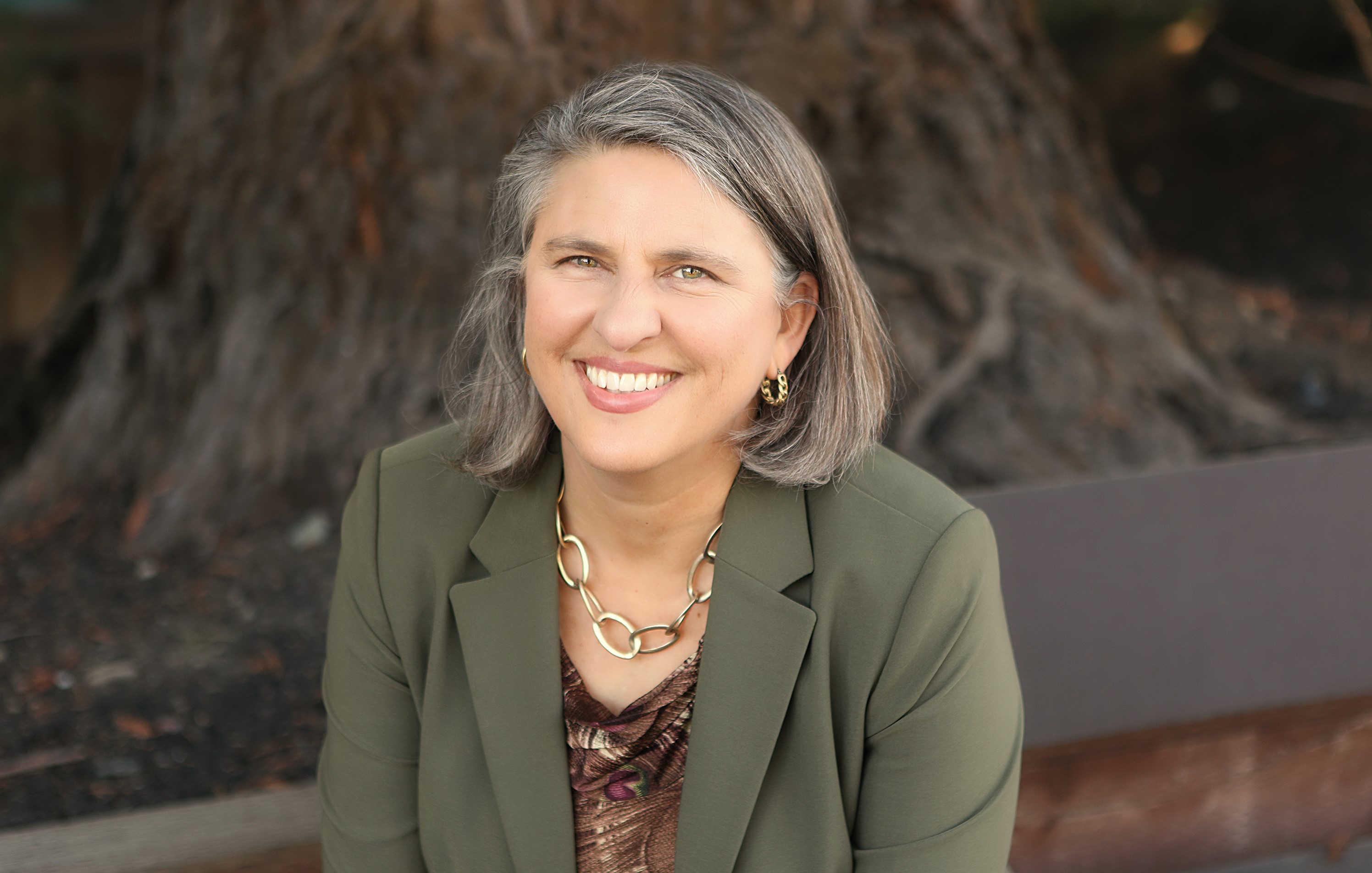 Edie Sparks smiles outdoors wearing a green jacket in front of a large tree