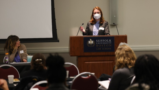 Attorney Eva Jellison, masked, speaks at a podium in front of a PowerPoint presentation in a crowded room