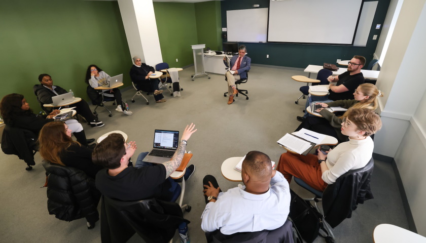 Ten students and Professor Rodrigues sit in a Suffolk classroom, desks arranged in a circle, as one student makes a point while gesturing