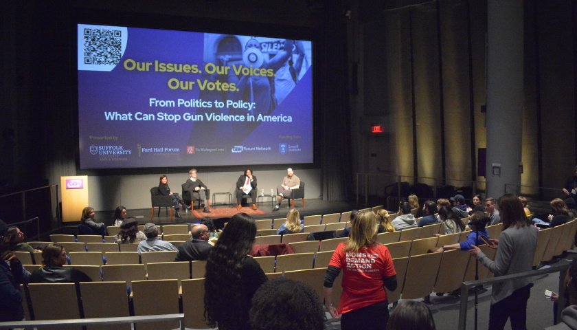 Several attendees line up to ask questions at a microphone in an auditorium as speakers sit in front of a screen with graphics and the event's title: "Our Issues. Our Voices. Our Votes. From Politics to Policy: What can stop gun violence in America?"