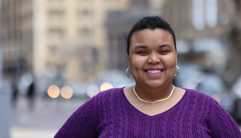 Rae'Niqua Victorine smiles as she poses outside of Suffolk University's 73 Tremont building in a purple sweater