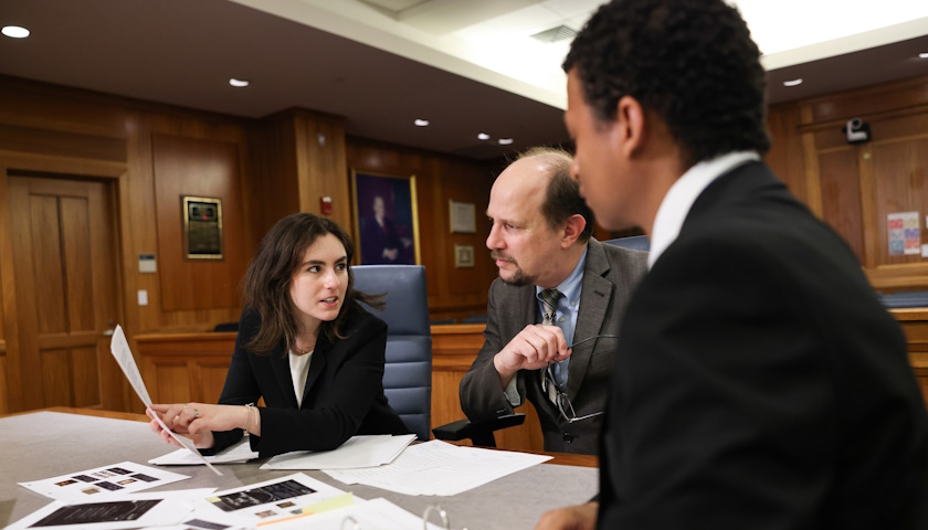 Brenna Koppel points to pieces of evidence as she, Professor Greg Bordelon, and Stephen Jimenez sit at a table in one of Suffolk’s mock court rooms