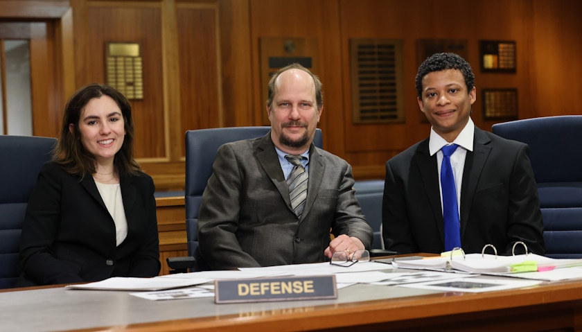 Brenna Koppel, Coach Greg Bordelon, and Stephen Jimenez, sit at a table labeled “defense” in one of Suffolk’s mock court rooms