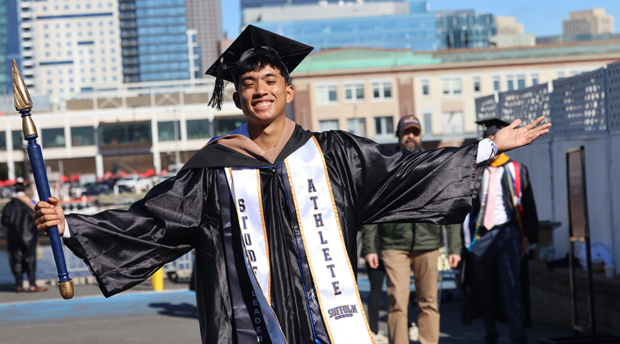A graduating student in regalia wear a student athlete stole stands with arms spread wide at Commencement 2025