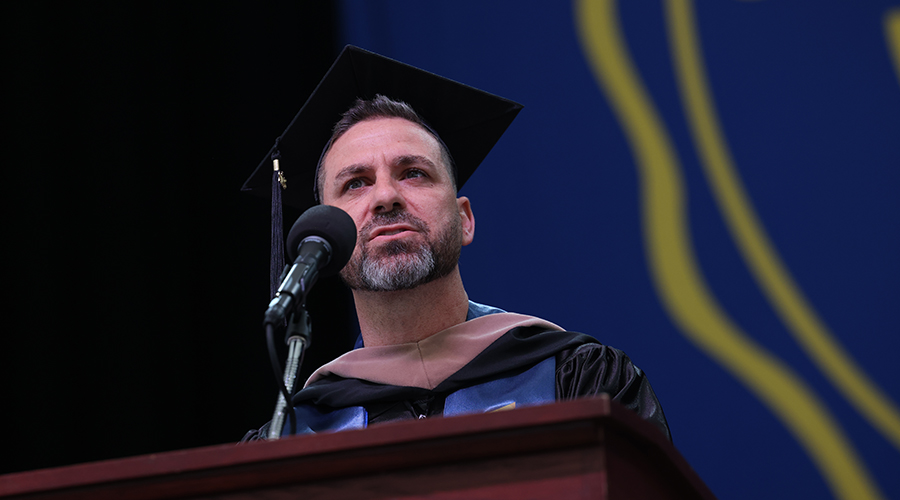 A man named Derek Goodwin speaking at the podium of the 2025 Sawyer Business School Commencement
