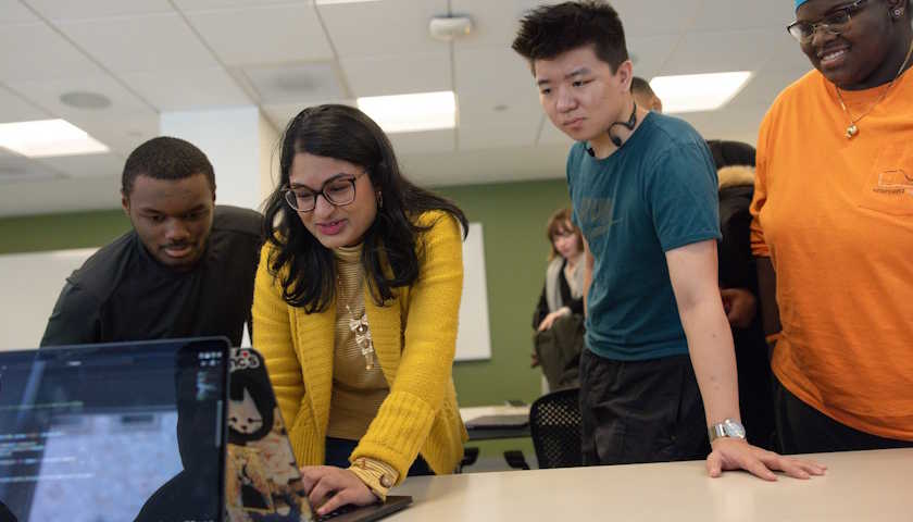 Four people crowd around two laptops in a classroom