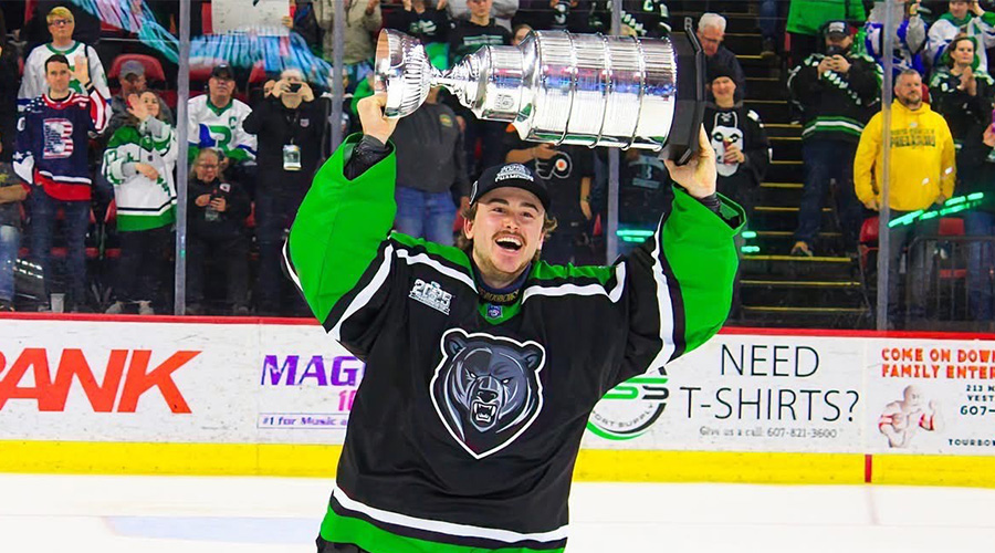 A Suffolk men's hockey player in a black and green jersey, skating on the ice, smiling and hoisting a trophy