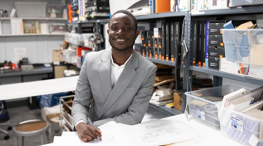 Adeleke Eyeowa, in a gray blazer, smiles as he writes at a table in Suffolk's interior architecture and design materials library