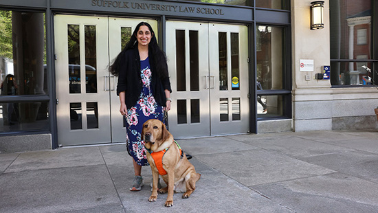 A woman, Rebecca Arrowood, and her guide dog in an orange vest, stand in front of the Suffolk University Law School building.